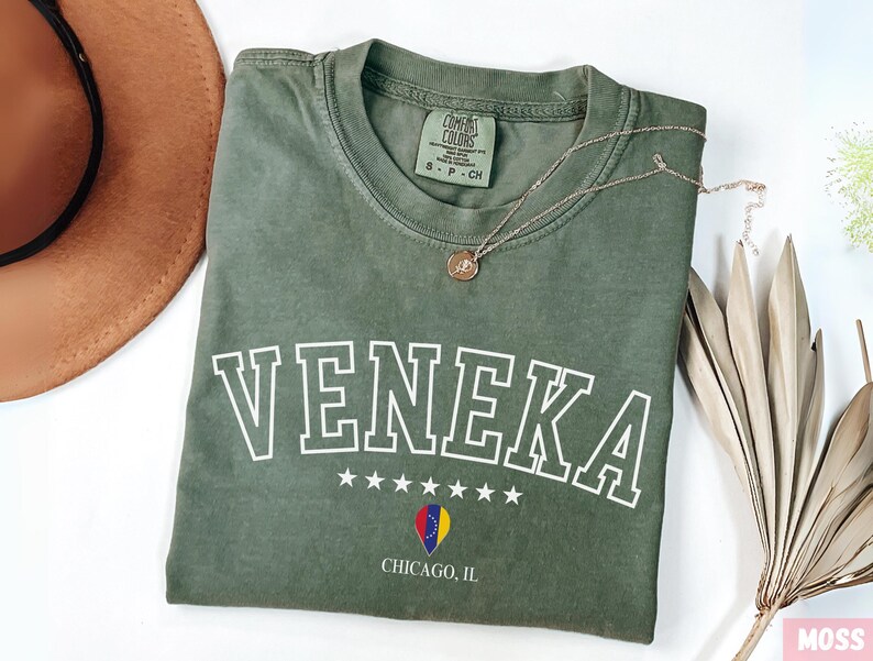 May include: A green t-shirt with the word "VENEKA" in white outlined letters, five white stars, and a small graphic of a flag. The text "CHICAGO, IL" is printed below the graphic. The t-shirt is folded and laid on a white surface.