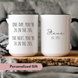 May include: Two white ceramic mugs with black handles and black rims. One mug has the text "ONE DAY, YOU'RE 20 IN THE 70S. THE NEXT, YOU'RE 70 IN THE 20S." The other mug has the text "Steve EST. 1955".