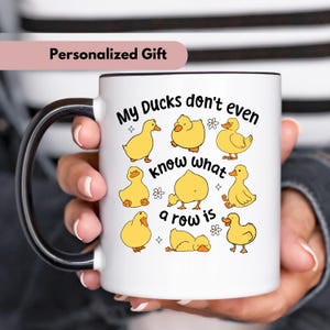 May include: White ceramic mug with a black handle and rim, featuring the text "My Ducks don't even know what a row is" and illustrations of yellow cartoon ducks. The mug is labeled "Personalized Gift".