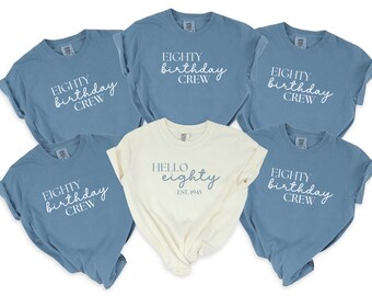 May include: A collection of blue and cream-colored t-shirts. The blue shirts display "EIGHTY birthday CREW" in white script. The cream shirt reads "HELLO eighty EST. 1945" in a similar font. A casual, celebratory theme.