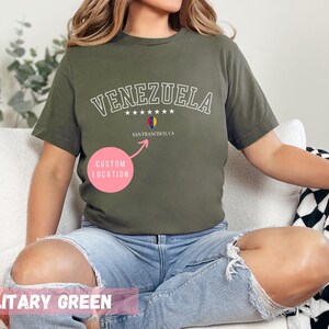 May include: Military green t-shirt with the word "VENEZUELA" in white, arched text above a small graphic of the Venezuelan flag and the text "SAN FRANCISCO, CA". The shirt is a solid color and has a crew neck.