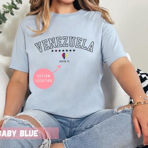 May include: Baby blue t-shirt with the word "VENEZUELA" in black, arched text. Below the text are six black stars and a graphic of the Venezuelan flag. The text "Miami, FL" is below the flag graphic. The words "CUSTOM LOCATION" are in a pink circle.