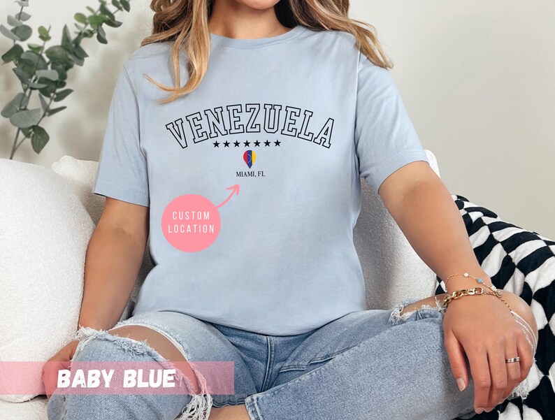 May include: Baby blue t-shirt with the word "VENEZUELA" in black, arched text. Below the text are six black stars and a graphic of the Venezuelan flag. The text "Miami, FL" is below the flag graphic. The words "CUSTOM LOCATION" are in a pink circle.