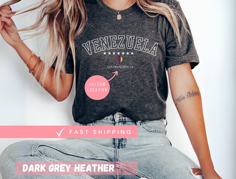May include: Dark grey heather t-shirt with the word "VENEZUELA" in white arched lettering. Below the word "VENEZUELA" are five white stars, a graphic, and the text "SAN FRANCISCO, CA". The shirt also has the text "CUSTOM LOCATION".