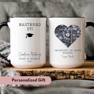 May include: Two white ceramic mugs with black handles and rims. One mug says "MASTERED IT!" with a graduation cap and the name "Carofinee Martinez" and "MASTER IN PHILOSOPHY -2026-". The other mug has a heart-shaped map and "UNIVERSITY OF SOUTH FLORIDA Tampa, Florida".