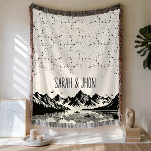 May include: Cream-colored woven throw blanket with a black constellation pattern at the top and a mountain and lake scene at the bottom. The names "SARAH & JHON" are printed in black. The blanket has a fringed edge.