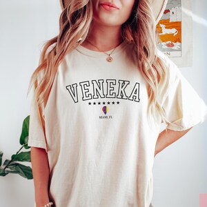 May include: A woman wearing a cream coloured t-shirt with the text "VENEKA" in black lettering, with five stars underneath the text and "MIAMI, FL" underneath the stars. The shirt has a small heart with the colours of the Miami flag.