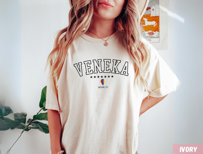 May include: A woman wearing a cream coloured t-shirt with the text "VENEKA" in black lettering, with five stars underneath the text and "MIAMI, FL" underneath the stars. The shirt has a small heart with the colours of the Miami flag.