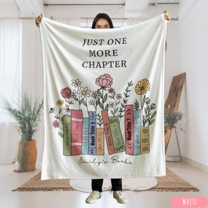 May include: A white blanket with the text "JUST ONE MORE CHAPTER" in black. The blanket features a design of colorful books with flowers and the text "Emily's Books". The blanket is white.