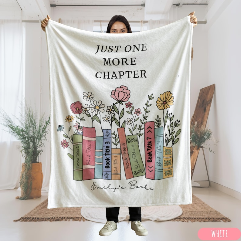 May include: A white blanket with the text "JUST ONE MORE CHAPTER" in black. The blanket features a design of colorful books with flowers and the text "Emily's Books". The blanket is white.