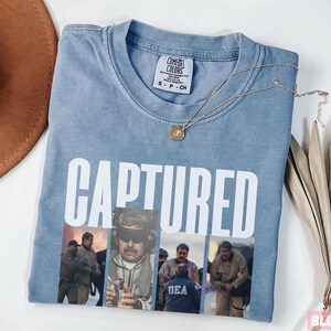 May include: Light blue t-shirt with the word "CAPTURED" in white block letters. Below the text is a collage of images. A gold necklace with a pendant rests on the shirt. A brown hat is visible in the upper left corner.
