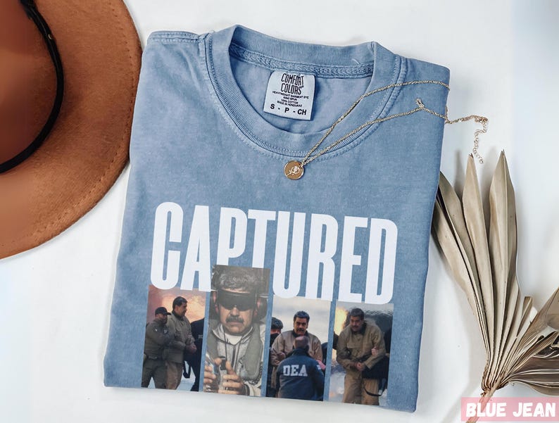 May include: Light blue t-shirt with the word "CAPTURED" in white block letters. Below the text is a collage of images. A gold necklace with a pendant rests on the shirt. A brown hat is visible in the upper left corner.