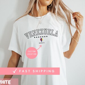 May include: White t-shirt with the word "VENEZUELA" in black, arched text above five stars. Below, a graphic of a flag and the text "MIAMI, FL". A pink circle with "CUSTOM LOCATION" is below the graphic.