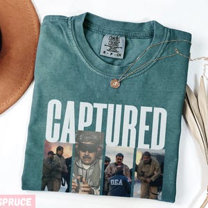 May include: Blue spruce t-shirt with the word "CAPTURED" in white block letters. The shirt features a collage of images of people in uniform. A gold necklace with a pendant rests on the shirt. A brown hat is visible in the upper left corner.