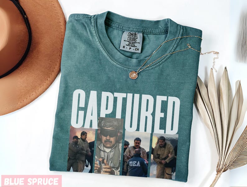 May include: Blue spruce t-shirt with the word "CAPTURED" in white block letters. The shirt features a collage of images of people in uniform. A gold necklace with a pendant rests on the shirt. A brown hat is visible in the upper left corner.