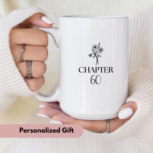May include: White ceramic mug with a black floral design and the text "CHAPTER 60". The mug has a large handle and is held by a person wearing a white sweater. The words "Personalized Gift" are at the bottom of the image.