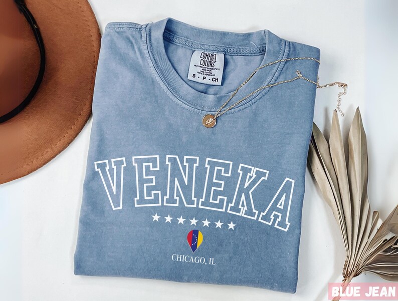 May include: A light blue t-shirt with the word "Veneka" printed in white with a white outline. The shirt also has a small graphic of a heart with the flag of Romania inside and the text "Chicago, IL" printed below.