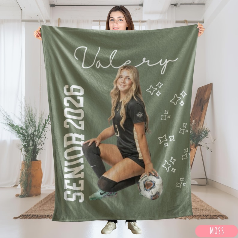 May include: A moss green blanket with the name "Valery" in white script and the text "SENIOR 2026" in white block letters. The blanket features a photo of a young woman in a soccer uniform holding a soccer ball. White star graphics are scattered across the blanket.