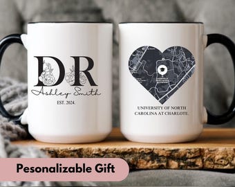 May include: Two white ceramic mugs with black handles and rims. One mug features "DR" with floral accents, the name "Ashley Smith", and "EST. 2024." The other mug displays a heart-shaped map of the University of North Carolina at Charlotte.