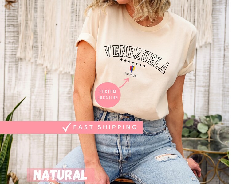 May include: Cream-colored t-shirt with the word "VENEZUELA" in black, arched lettering. Below the word are five black stars and a graphic of a colorful hot air balloon with the text "MIAMI, FL". A pink circle with "CUSTOM LOCATION" is also visible.