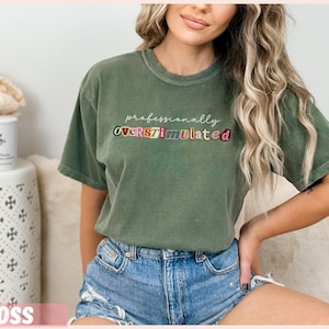 May include: A moss green t-shirt featuring the text "professionally OVERSTIMULATED" in colourful letters. The shirt has a crew neck and short sleeves. The model is wearing denim shorts.