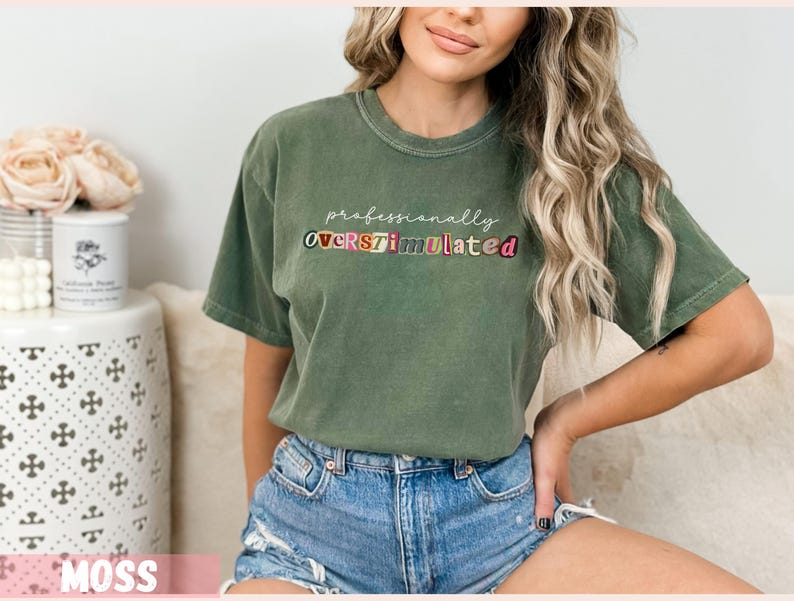 May include: A moss green t-shirt featuring the text "professionally OVERSTIMULATED" in colourful letters. The shirt has a crew neck and short sleeves. The model is wearing denim shorts.