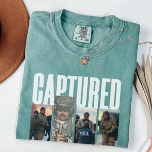 May include: A light green t-shirt with the word "CAPTURED" in large white letters. Below the text are four images of people. A gold necklace with a pendant is on the shirt. A brown hat and a decorative plant are also in the image.