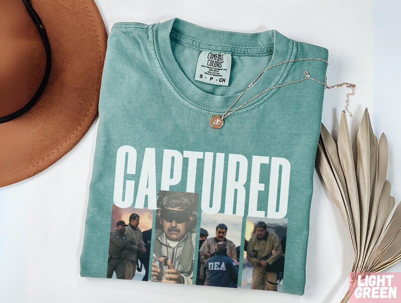 May include: A light green t-shirt with the word "CAPTURED" in large white letters. Below the text are four images of people. A gold necklace with a pendant is on the shirt. A brown hat and a decorative plant are also in the image.