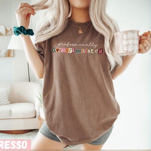 May include: A brown t-shirt with the text "professionally OVERSTIMULATED" in colourful letters. The person is wearing a green scrunchie and holding a pink mug with the words "cool mum vibes" on it.