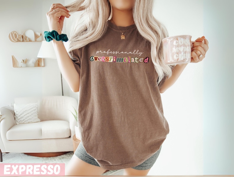 May include: A brown t-shirt with the text "professionally OVERSTIMULATED" in colourful letters. The person is wearing a green scrunchie and holding a pink mug with the words "cool mum vibes" on it.