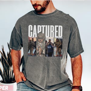 May include: A gray t-shirt with the word "CAPTURED" in white block letters. Below the text are three images of people. The shirt has a vintage, washed look. The person in the image is wearing a watch.
