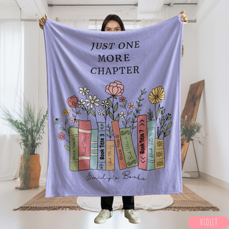May include: A violet-colored blanket with the text "JUST ONE MORE CHAPTER" and a floral design. The design includes colorful books with titles and flowers. The blanket also has the text "Emily's Books".