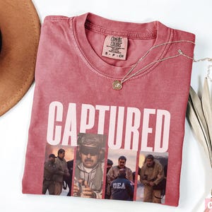 May include: Crimson-colored t-shirt with the word "CAPTURED" in white block letters. Below the text are four images of people. A gold-colored necklace with a pendant rests on the shirt. A brown hat and dried plant are in the background.