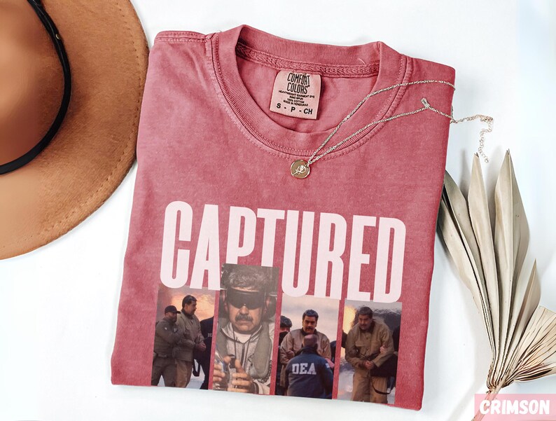 May include: Crimson-colored t-shirt with the word "CAPTURED" in white block letters. Below the text are four images of people. A gold-colored necklace with a pendant rests on the shirt. A brown hat and dried plant are in the background.