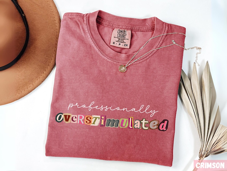 May include: Crimson t-shirt with the words "professionally overstimulated" in various fonts and colours. A gold necklace with a pendant lies on the shirt. A brown hat and dried plant are in the background.