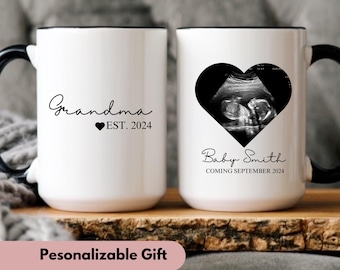 May include: Two white coffee mugs with black handles and black rims. The mug on the left says "Grandma EST. 2024". The mug on the right has a black heart with a sonogram image inside and says "Baby Smith Coming September 2024".