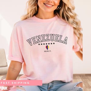 May include: A light pink t-shirt with the word "VENEZUELA" arched across the top in black letters. Below the word are five black stars and a graphic of a hot air balloon. The text "MIAMI, FL" is below the balloon.