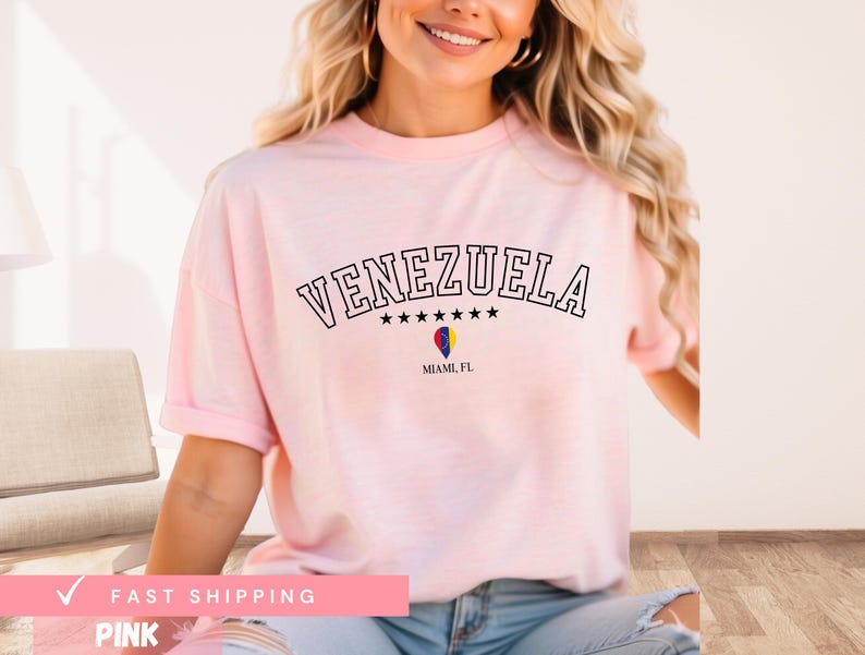 May include: A light pink t-shirt with the word "VENEZUELA" arched across the top in black letters. Below the word are five black stars and a graphic of a hot air balloon. The text "MIAMI, FL" is below the balloon.