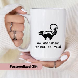 May include: White ceramic mug with a black and white skunk illustration wearing a graduation cap. The mug has the text "so stinking proud of you!" printed in black. The mug is a personalized gift.