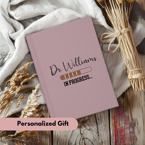 Pode incluir: Um caderno rosa personalizado com o texto "Dr. Williams IN PROGRESS..." e um gr&aacute;fico de barra de progresso. Presente personalizado.