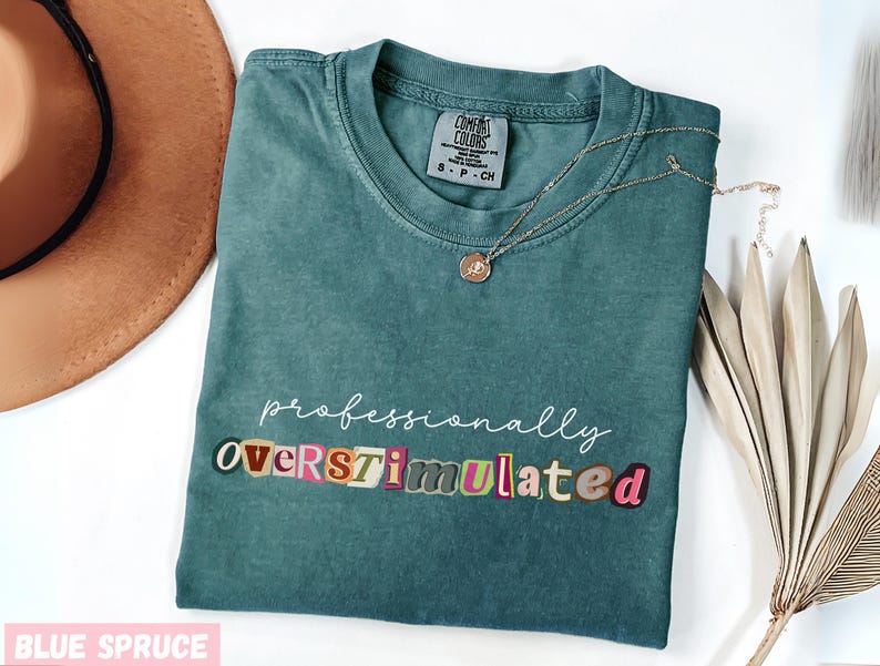 May include: Teal blue t-shirt with the text "professionally overstimulated" in a colourful, block letter design. A gold necklace with a pendant is on the shirt. A brown hat and decorative leaves are also in the image.