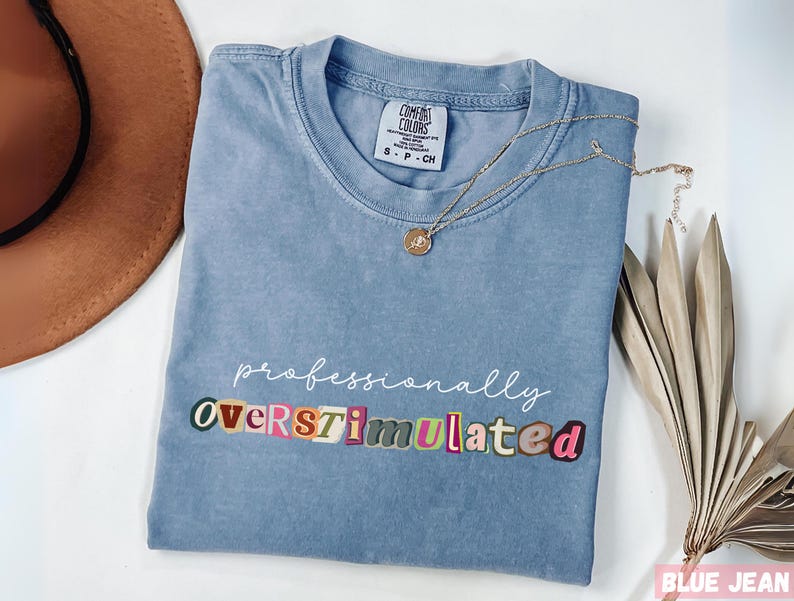 Folded blue Comfort Colors® t-shirt featuring professionally in script above colorful cutout style lettering spelling overstimulated across the front.