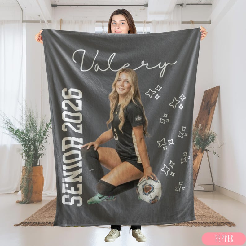 May include: A dark gray blanket featuring a photo of a young woman in a soccer uniform, holding a soccer ball. The blanket has the name "Valery" in cursive and the text "Senior 2026". White star graphics are scattered across the blanket.