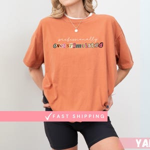May include: An orange t-shirt with the text "professionally overstimulated" in various colours. The model is wearing black shorts and a necklace. The shirt has short sleeves.