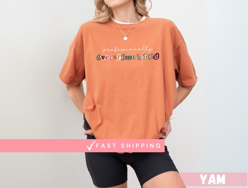May include: An orange t-shirt with the text "professionally overstimulated" in various colours. The model is wearing black shorts and a necklace. The shirt has short sleeves.