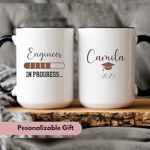May include: Two white ceramic mugs with black rims. The mug on the left has the text "Engineer" with a progress bar that says "In Progress..." The mug on the right has the text "Camila -2025- " with a graduation cap.