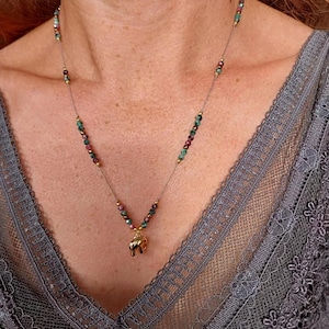 Peut inclure: Un collier délicat avec un pendentif éléphant doré. Le collier est orné de petites perles colorées dans les tons de vert, de rouge et d'or. Le collier est porté sur un haut en dentelle grise.