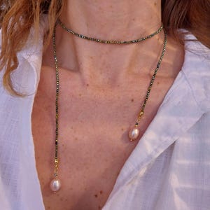 Puede incluir: Un collar de dos capas con un choker y una hebra más larga. El choker está hecho de pequeñas cuentas iridiscentes. La hebra más larga tiene un diseño similar con cuentas doradas y una perla rosa pálido colgante. El collar se lleva sobre una camisa blanca.
