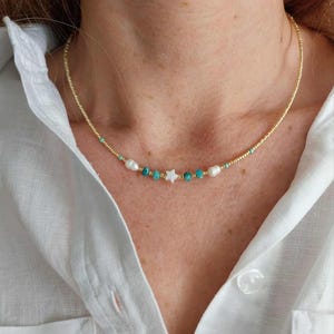 Può includere: Delicata collana d'oro con un piccolo ciondolo a forma di stella bianca, perle d'acqua dolce e perline turchesi. La collana è infilata su una sottile catena d'oro. La collana è indossata su una camicia bianca abbottonata.