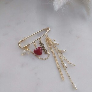 May include: Gold-coloured safety pin brooch with dangling chains, faux pearls, and a red heart charm. The brooch also features the letters "AMA" in gold. A stylish accessory.